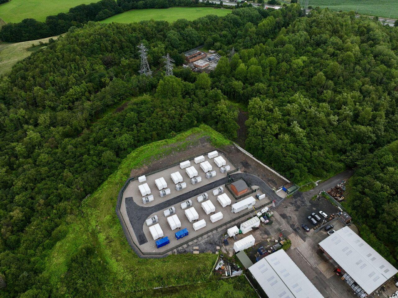 Aerial view of a battery energy storage site: rows of BESS containers on a hillside platform beside woodland.